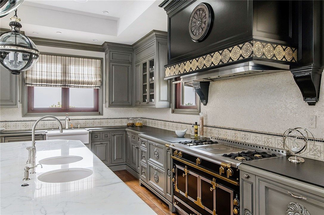 This elegant kitchen features dark grey cabinetry paired with a large, light-toned stone island and a prominent, ornate black range hood. The space is accented by decorative tile work along the backsplash and a sophisticated pendant light fixture hanging above the island. The perspective captures the functional layout of the cooking area, highlighting the high-end appliances and classic, detailed cabinetry design.
