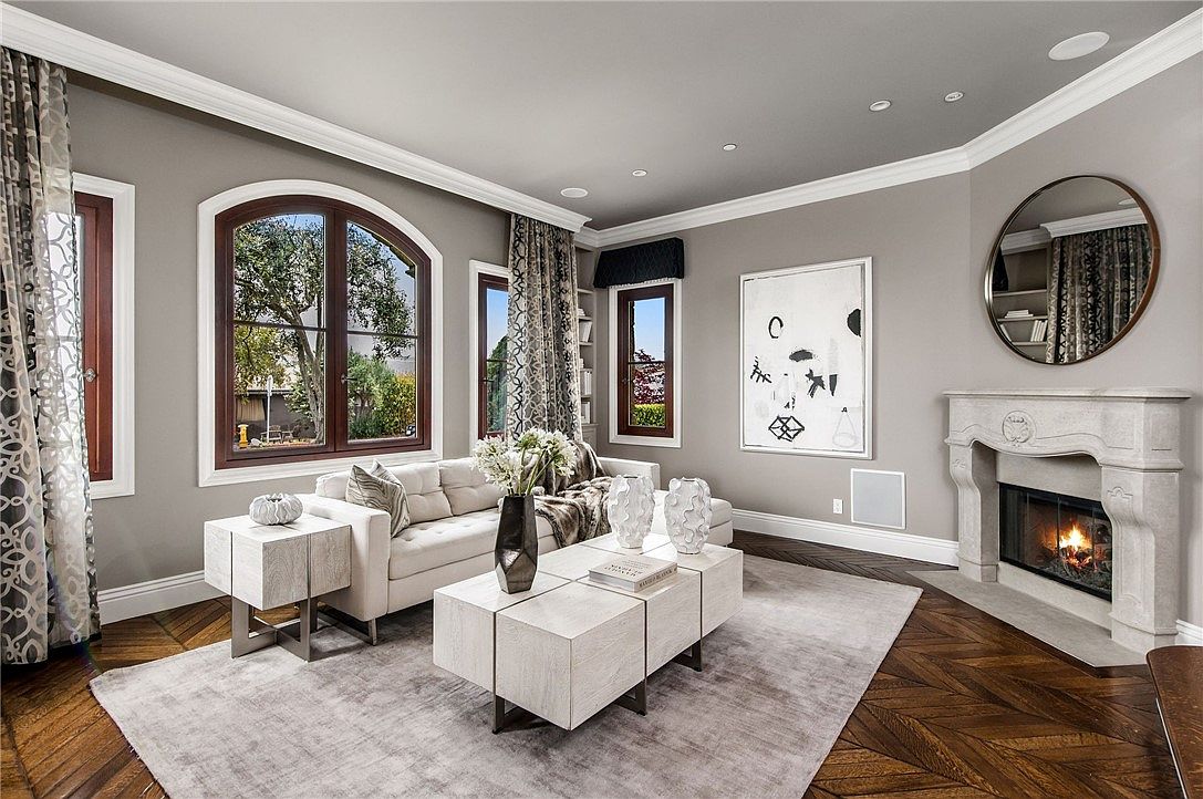 This elegant living room features a sophisticated neutral color palette, highlighted by a plush light-colored sofa and a modern, multi-sectioned coffee table. The space is anchored by a stone fireplace with a classic mantel and a circular mirror above, while rich, dark wood floors in a herringbone pattern provide a warm contrast. Large, arched windows dressed in patterned drapery allow natural light to fill the room, creating a refined and inviting atmosphere.