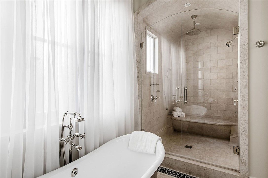 This elegant primary bathroom features a freestanding soaking tub positioned next to a large window draped in soft, sheer curtains. Adjacent to the tub is a spacious walk-in shower enclosed by glass, showcasing stone-tiled walls, a built-in stone bench, and dual shower heads. The space exudes a serene and luxurious atmosphere, highlighted by polished chrome fixtures and a neutral color palette.
