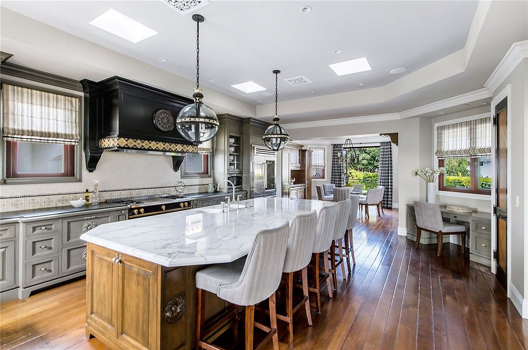 This spacious, high-end kitchen features a large central island with a stone countertop and seating for five, serving as the focal point of the room. The space is defined by a mix of dark cabinetry, a prominent black range hood, and warm wood flooring, all illuminated by recessed lighting and two large spherical pendant lights. A dedicated desk area is tucked into a windowed alcove, while an open-concept layout provides a clear view into the adjacent dining area.