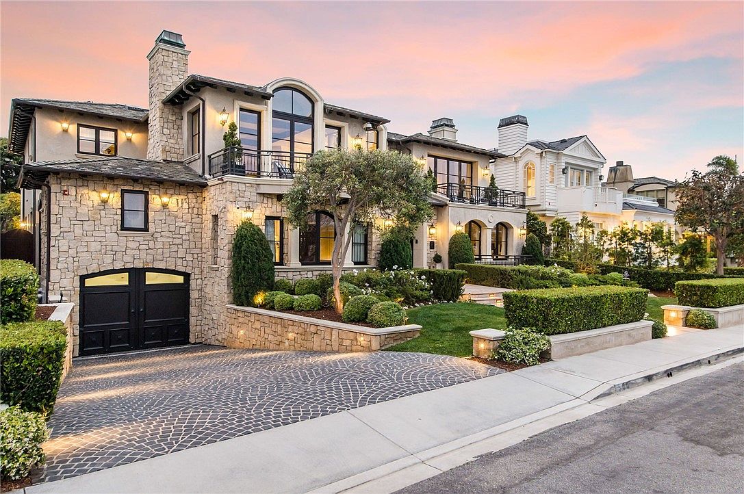 This elegant multi-story residence features a sophisticated stone facade with a prominent arched window and a private balcony. The front yard is meticulously landscaped with manicured hedges and a central tree, while a patterned stone driveway leads to a dark-toned garage. Captured at dusk, the warm exterior lighting creates an inviting and luxurious atmosphere.