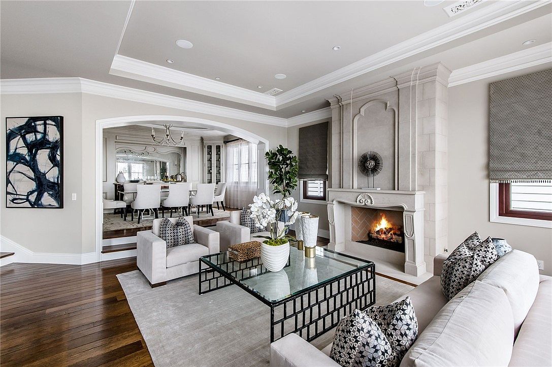 This elegant living room features a sophisticated neutral color palette, centered around a prominent stone fireplace with a tall, decorative surround. A glass-topped coffee table sits on a plush area rug, flanked by comfortable light-toned seating and patterned accent pillows. The space flows seamlessly into an adjacent dining area through a wide, arched opening, creating an open and airy atmosphere suitable for entertaining.