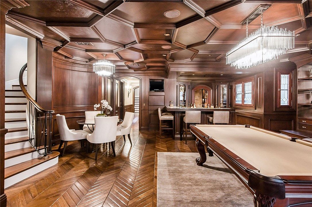 This sophisticated basement recreation area features rich wood-paneled walls and a complex coffered ceiling, creating a warm and luxurious atmosphere. The space includes a billiards area with a pool table, a dedicated dining nook with a circular table, and a built-in bar with stools. The room is finished with dark wood flooring laid in a herringbone pattern, providing a cohesive and high-end aesthetic.
