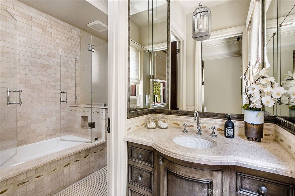 This elegant bathroom features a vanity with a stone countertop and dark wood cabinetry, complemented by a large mirror and a decorative pendant light. To the left, a glass-enclosed shower area includes a built-in bathtub surrounded by light-toned, textured wall tiles. The space is well-lit and maintains a sophisticated, traditional aesthetic.