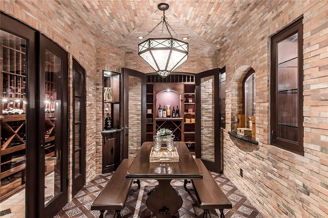 This sophisticated wine cellar features a barrel-vaulted brick ceiling and walls, creating an intimate, old-world atmosphere. A central wooden table with two matching benches serves as a tasting area, illuminated by a large, ornate geometric pendant light. The space is enclosed by dark-framed glass doors and includes built-in wine storage racks, showcasing a refined and luxurious design.