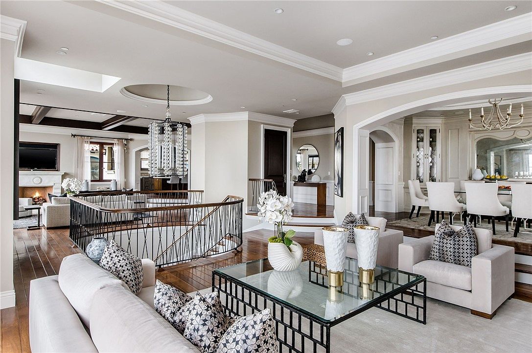 This expansive, open-concept living area features a sophisticated neutral color palette and high-end finishes, including a curved staircase with a decorative metal railing. The foreground showcases a contemporary glass-topped coffee table surrounded by plush seating, while the background reveals a formal dining space and a secondary living area with a fireplace. The perspective is from a slightly elevated vantage point, emphasizing the airy, interconnected layout and the elegant architectural details like the recessed ceiling and crystal chandelier.