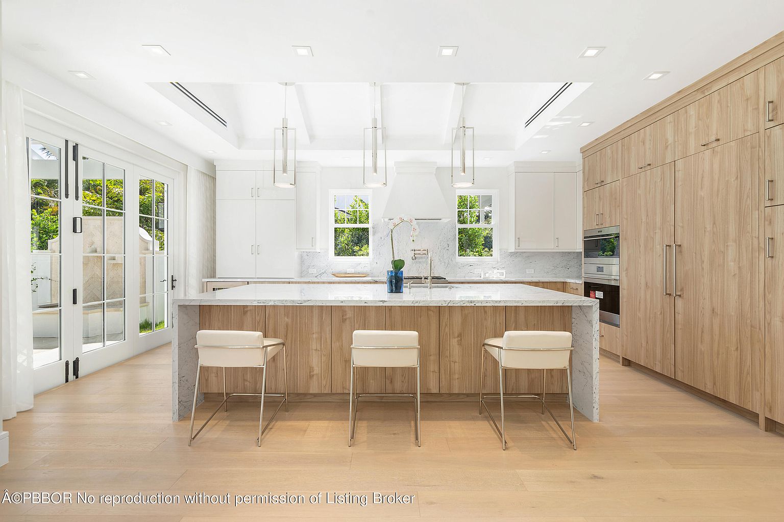 This is a bright and modern kitchen featuring a large island with seating, light wood cabinetry, and white countertops. The kitchen is well-lit with recessed lighting and pendant lights, and large windows provide natural light and views of the outdoors. The overall impression is clean, spacious, and luxurious.