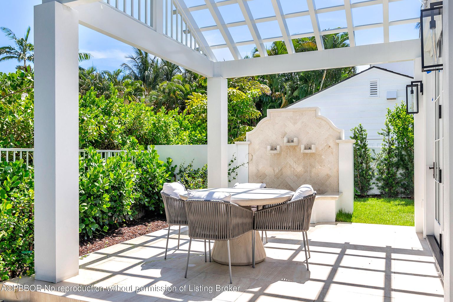 This image showcases a beautifully designed outdoor patio area, featuring a round table with four chairs under a white pergola. Lush greenery surrounds the space, creating a private and serene atmosphere. A decorative water feature adds a touch of elegance, making it an ideal spot for relaxation or entertaining.