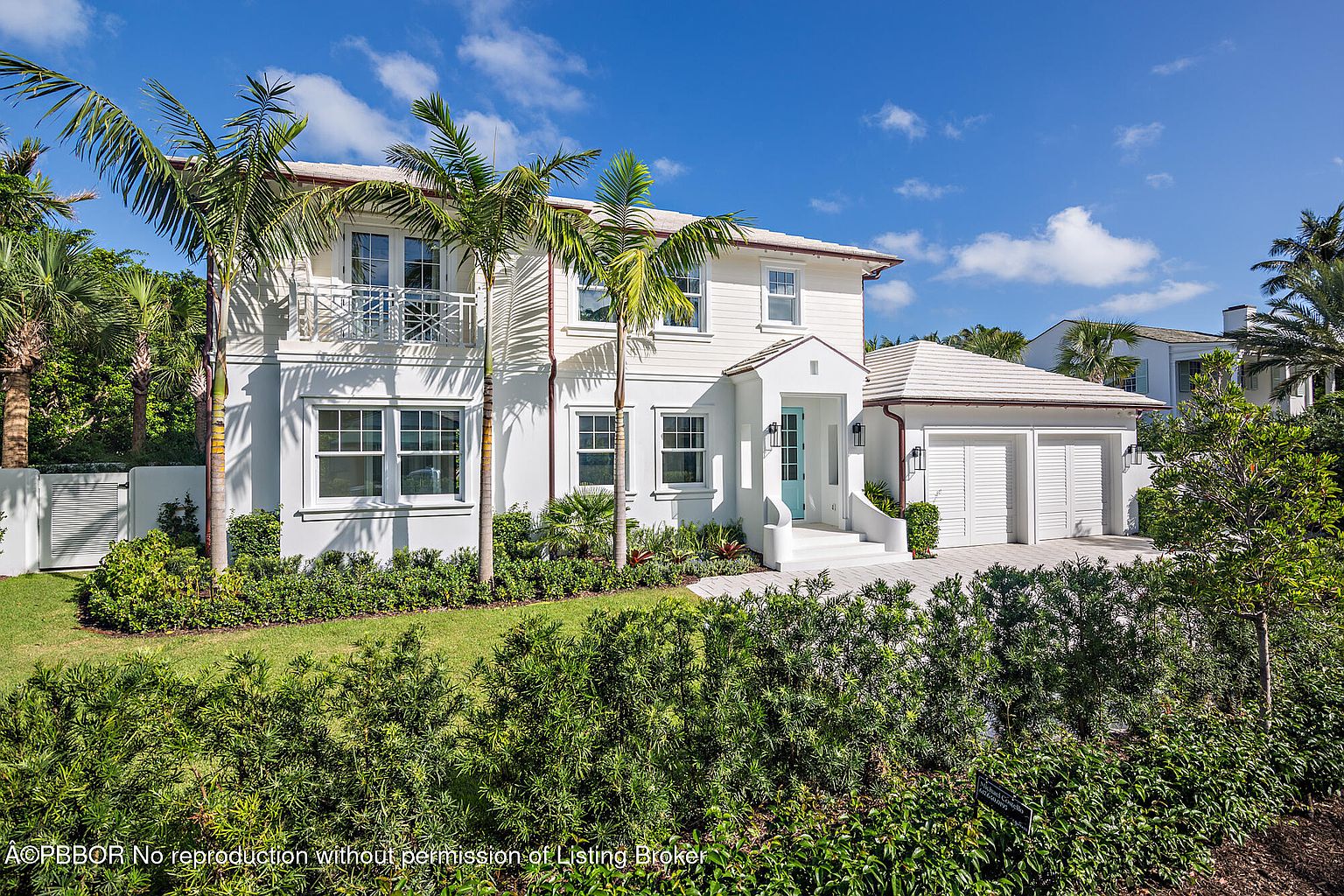 This is a front exterior view of a two-story white house with a detached garage. The house features a light blue front door, white trim, and a balcony on the second floor. Lush green landscaping surrounds the property, including palm trees and manicured bushes, creating a welcoming and upscale curb appeal.