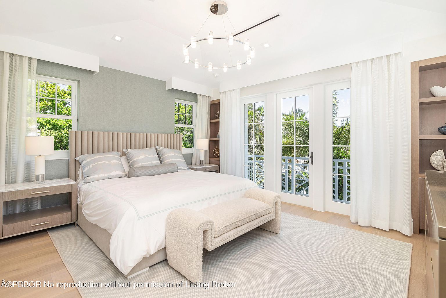 This is a bright and airy primary bedroom featuring a neutral color palette. The room includes a large bed with a padded headboard, matching nightstands, and a cozy bench at the foot of the bed. French doors lead to a balcony, providing ample natural light and a view of the surrounding greenery.