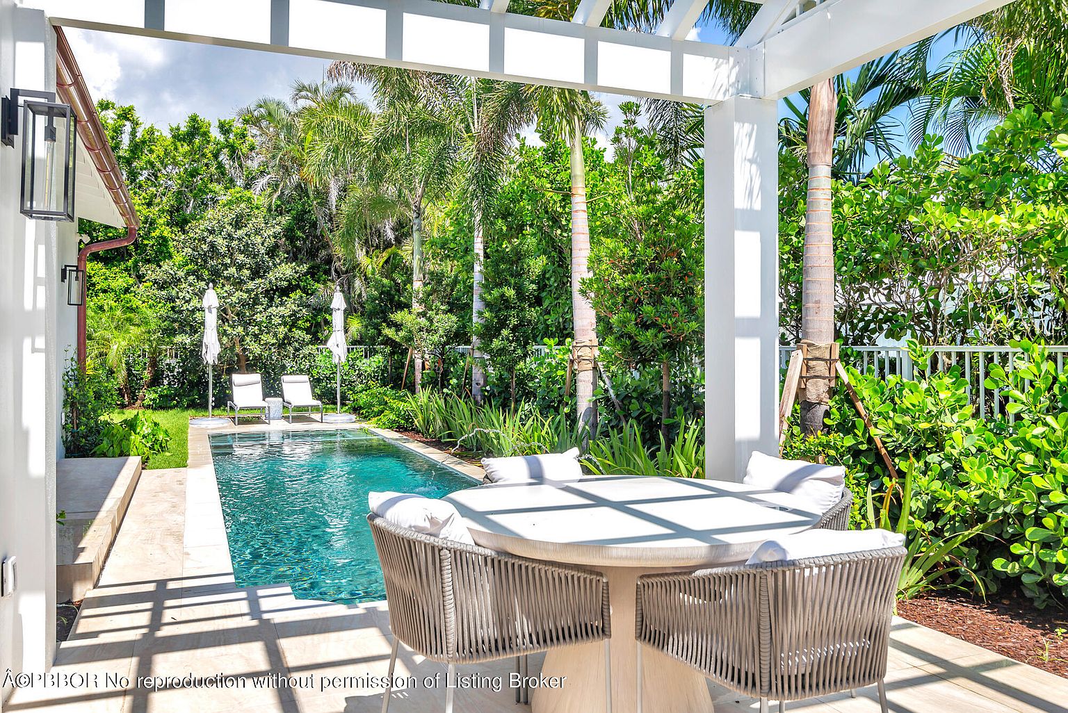 This image showcases a luxurious outdoor living space featuring a rectangular swimming pool surrounded by lush greenery and palm trees. A covered patio area with a round dining table and comfortable chairs provides a perfect spot for relaxation and entertaining. The overall impression is one of tranquility and upscale outdoor living.
