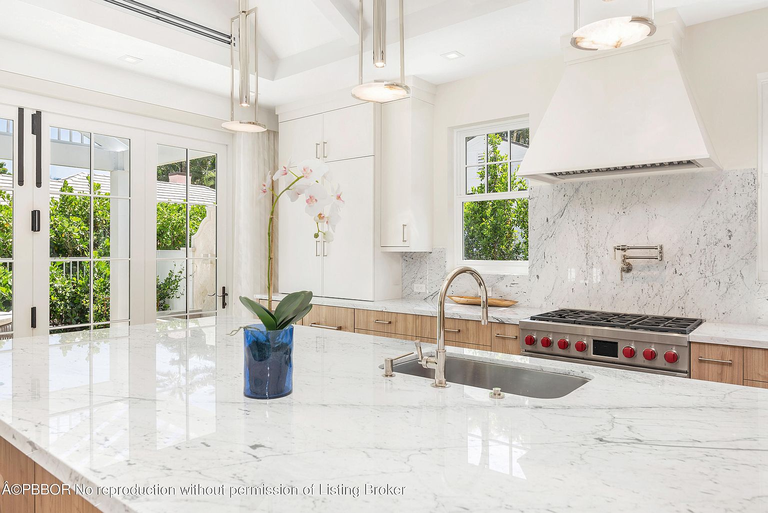 This is a bright and modern kitchen featuring a large marble island with a stainless steel sink and faucet. The kitchen includes white cabinetry with wood accents, a stainless steel range with a white hood, and marble backsplash. Large windows and doors provide natural light and a view to the outside, enhancing the open and airy feel of the space.