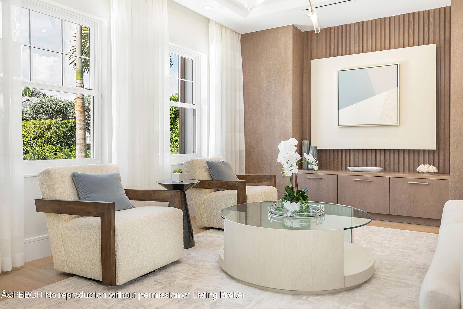 This is an interior shot of a living room featuring two armchairs with light-colored upholstery and wooden frames, positioned around a round glass-topped coffee table. Large windows with sheer white curtains provide natural light, and a built-in cabinet with artwork above it adds a touch of sophistication. The room is decorated in neutral tones, creating a bright and inviting atmosphere.