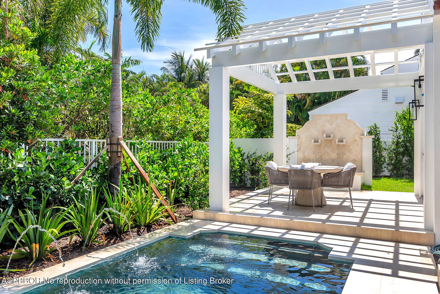 This image showcases a luxurious outdoor pool and spa area, featuring a modern pergola with a dining set, lush landscaping, and a serene water feature. The pool's clear blue water invites relaxation, while the surrounding greenery provides privacy and a tropical ambiance. The overall design exudes sophistication and tranquility, perfect for upscale outdoor living.