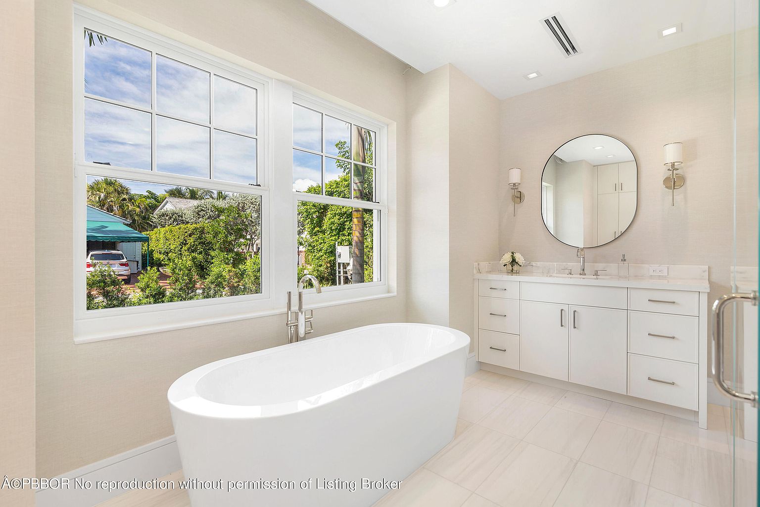 This is a bright and modern primary bathroom featuring a freestanding white bathtub positioned in front of two large windows offering a view of lush greenery. A white vanity with a marble countertop and an oval mirror are situated against a light beige textured wall, complemented by sconce lighting. The flooring consists of large, light-colored tiles, enhancing the spacious and clean aesthetic of the bathroom.