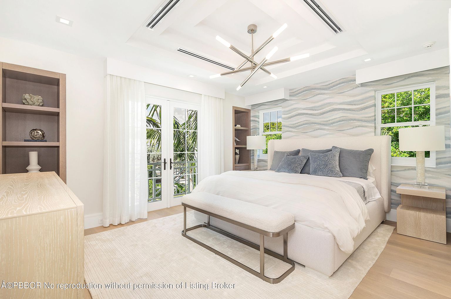 This is a bright and airy primary bedroom featuring a large bed with blue accent pillows, flanked by matching nightstands and lamps. French doors lead to a balcony, and the room is decorated in a modern, minimalist style with a neutral color palette. The room is well-lit with natural light and a modern chandelier.