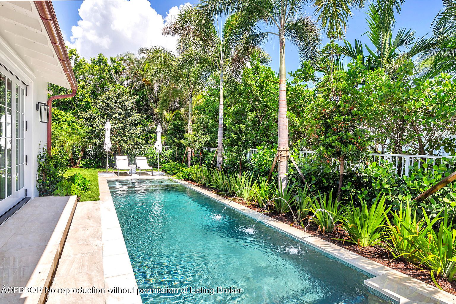 This image showcases a luxurious outdoor pool area, featuring a long, narrow pool with water features. Lush tropical landscaping surrounds the pool, providing privacy and a serene atmosphere. Two lounge chairs with umbrellas are placed at the end of the pool, inviting relaxation and leisure.