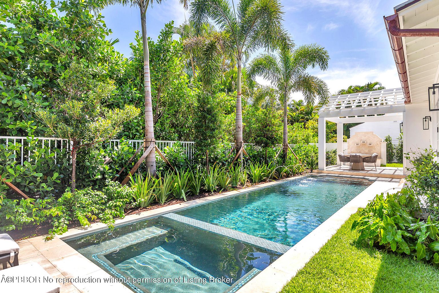 This image showcases a luxurious backyard pool and spa area, surrounded by lush tropical landscaping. The rectangular pool features a built-in spa with mosaic tile accents, and a white pergola with outdoor seating provides a relaxing lounge area. The scene evokes a sense of tranquility and upscale outdoor living.