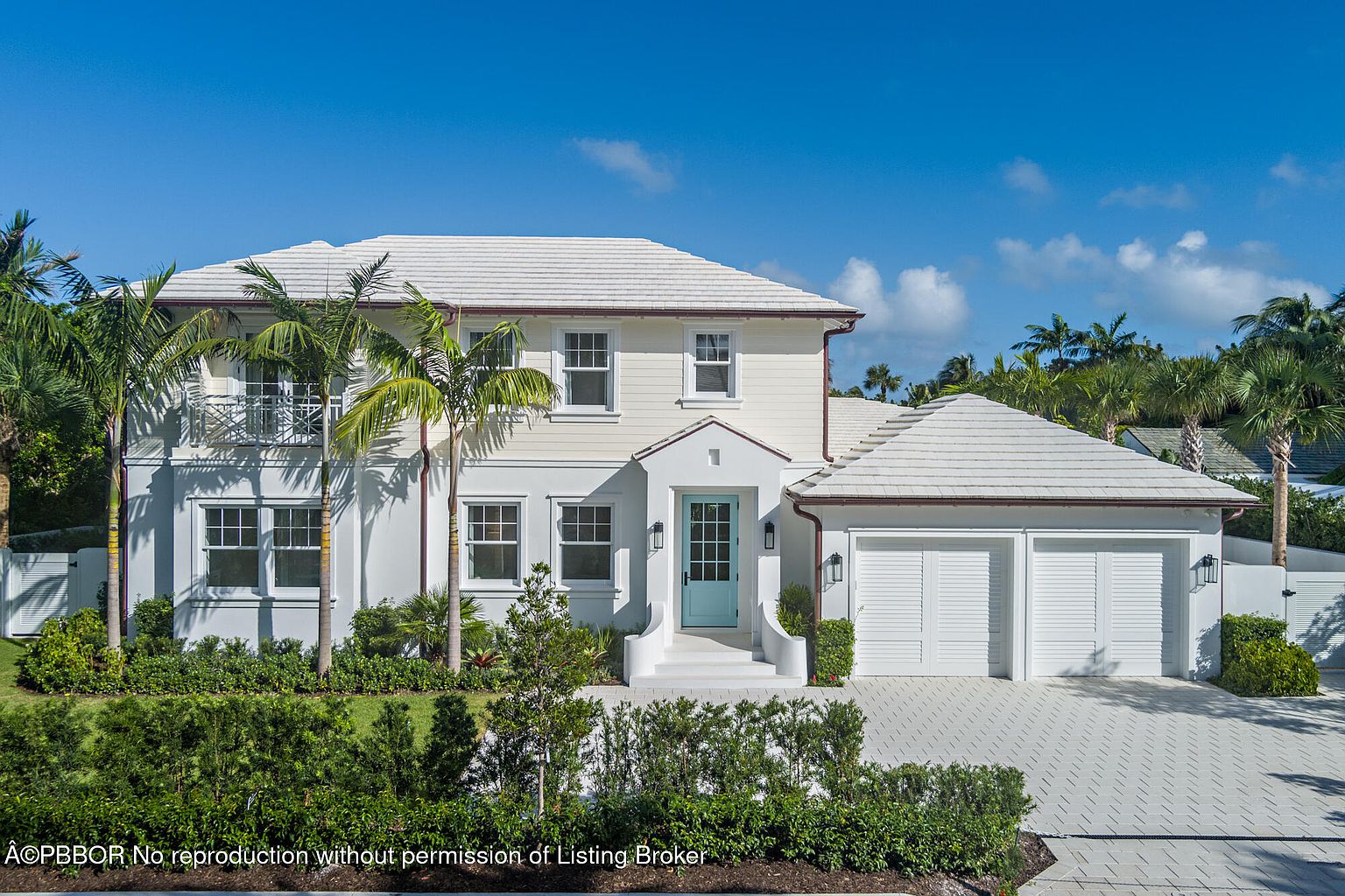 This is a front view of a two-story luxury home with a white exterior, a light-colored tile roof, and a striking turquoise front door. The property features well-manicured landscaping, including lush green hedges and palm trees, enhancing its curb appeal. A two-car garage is attached to the side of the house, and a paved driveway leads up to it.