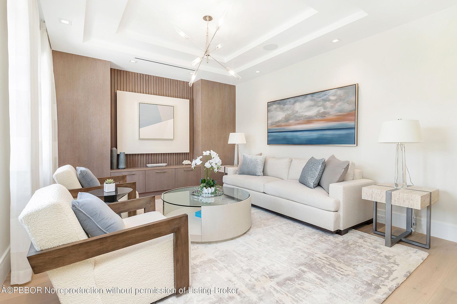 This is a bright and elegant living room featuring a neutral color palette with pops of blue in the artwork and pillows. The room is furnished with a light-colored sofa, two armchairs, and a round glass-topped coffee table. A large abstract painting hangs above the sofa, and a modern chandelier illuminates the space, creating a sophisticated and inviting atmosphere.