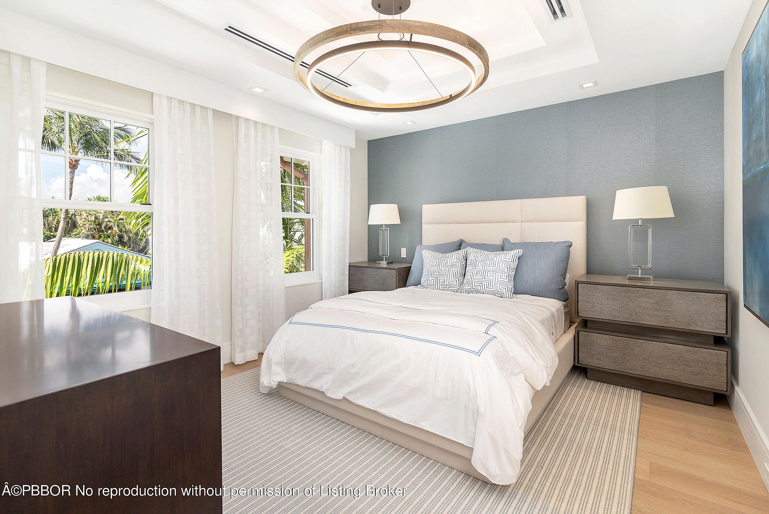 This is a bright and airy bedroom featuring a queen-sized bed with white linens and blue accents. The room has two large windows with sheer white curtains, allowing natural light to flood the space. A modern, circular chandelier hangs from the ceiling, and the walls are painted in a calming blue hue, creating a serene and inviting atmosphere.
