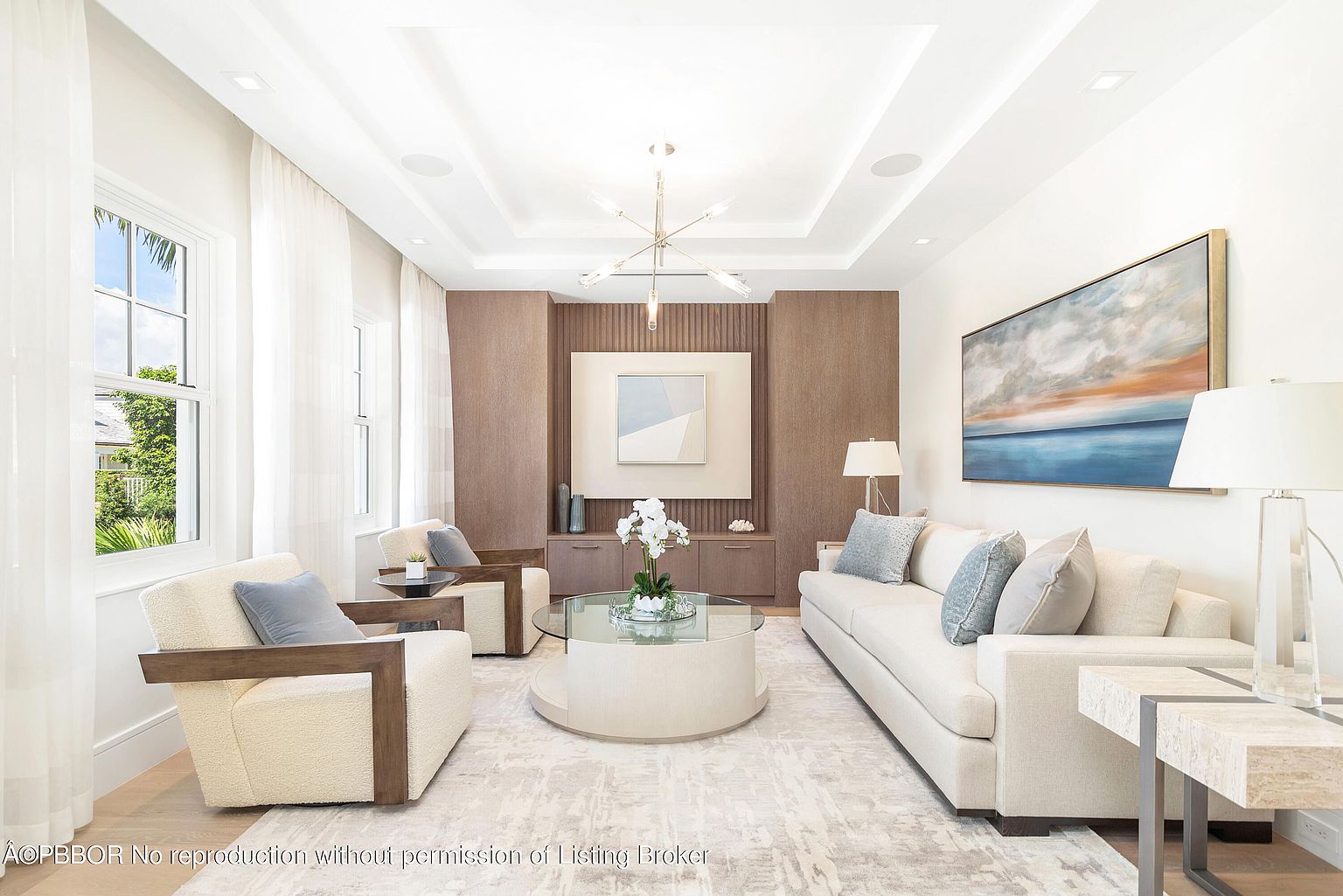This is a bright and elegant living room featuring a neutral color palette with pops of blue. The room is furnished with a white sofa, two armchairs, and a round glass coffee table. A large abstract painting hangs above the sofa, and natural light streams in through the windows, creating a welcoming and sophisticated atmosphere.