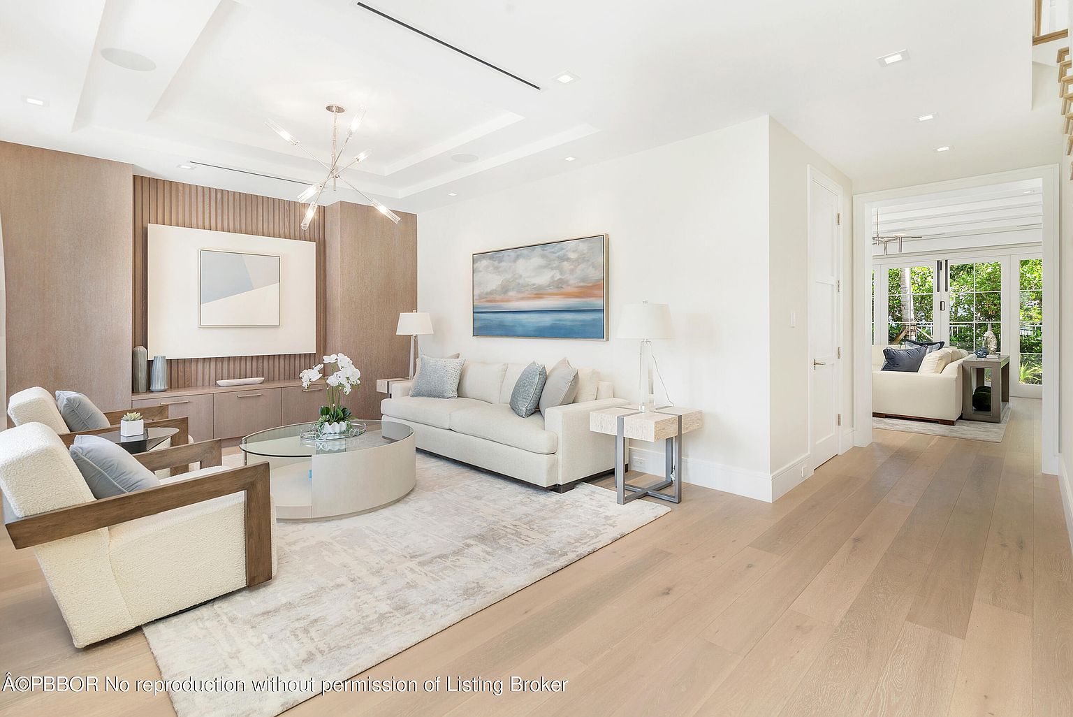 This is a bright and airy living room featuring a neutral color palette with light wood flooring and white walls. The room is furnished with a comfortable sofa, two armchairs, and a round glass-top coffee table. Artwork and decorative lighting add to the sophisticated ambiance, while a glimpse into an adjacent room suggests an open and inviting floor plan.