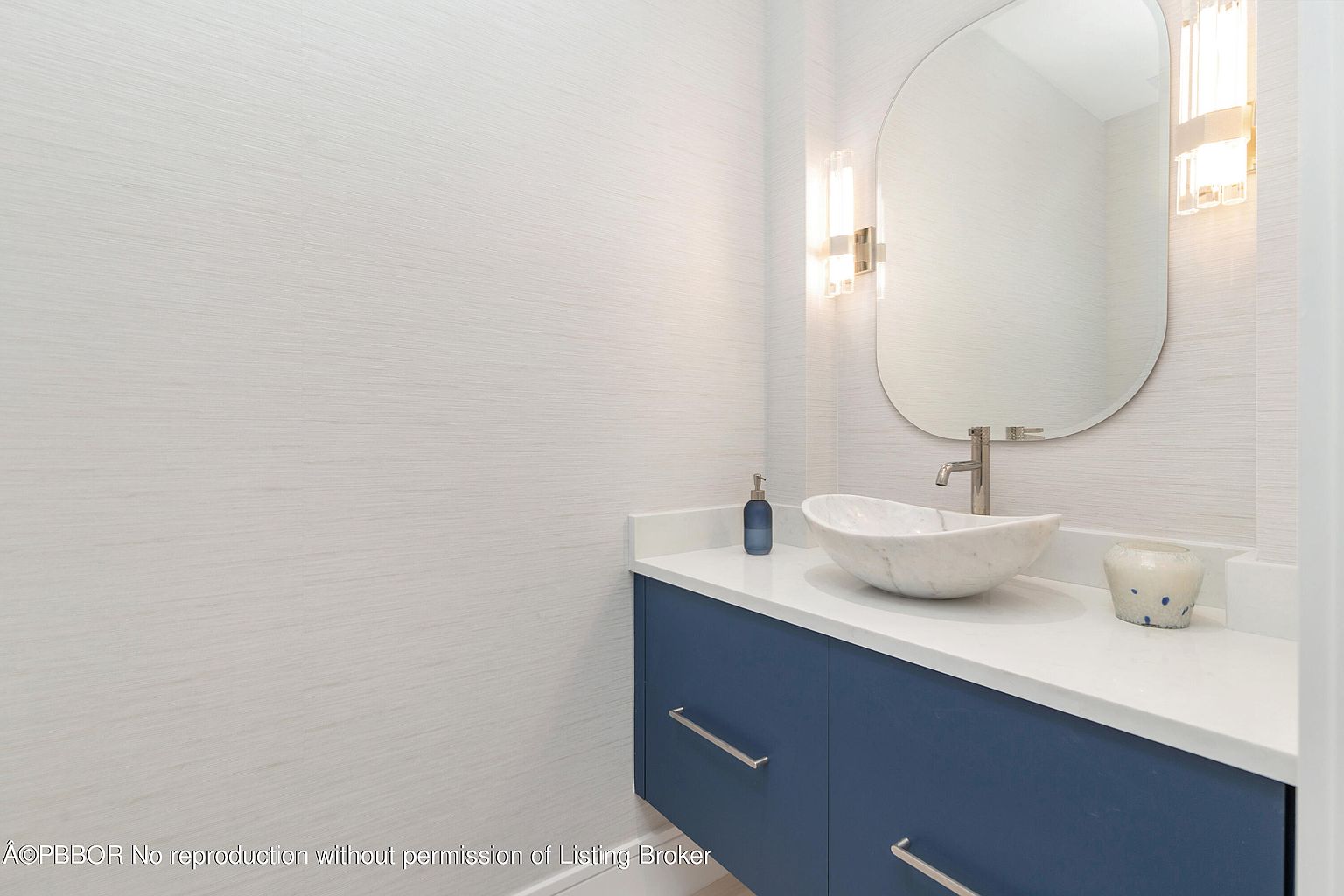 This is a well-lit guest bathroom featuring a modern design. The vanity has a blue cabinet with a white countertop, complemented by a vessel sink and a sleek faucet. An oval mirror hangs above the sink, flanked by contemporary sconces, creating a clean and stylish space.