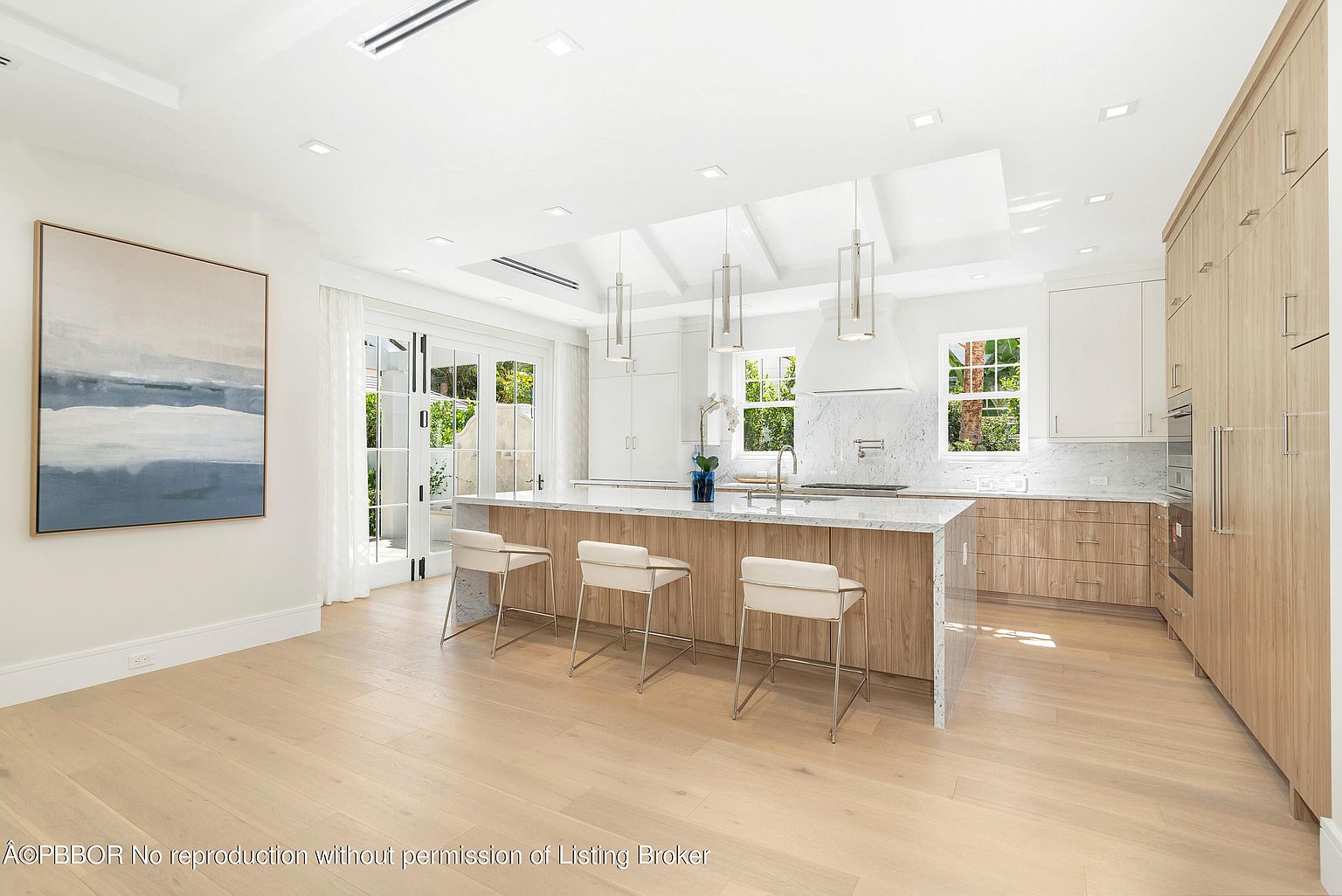 This is a bright and spacious kitchen featuring a large center island with seating, light wood cabinetry, and marble countertops. The kitchen is well-lit with recessed lighting and pendant lights over the island, and natural light streams in through the windows and glass doors. The overall impression is one of modern elegance and functionality.