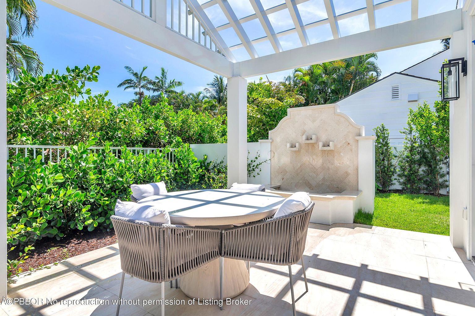 This image showcases a beautifully designed outdoor patio area, featuring a round dining table with woven chairs and plush cushions. The patio is covered by a white pergola, providing shade and an elegant architectural element. A decorative water fountain adds a touch of sophistication, while lush greenery in the background creates a serene and private atmosphere.