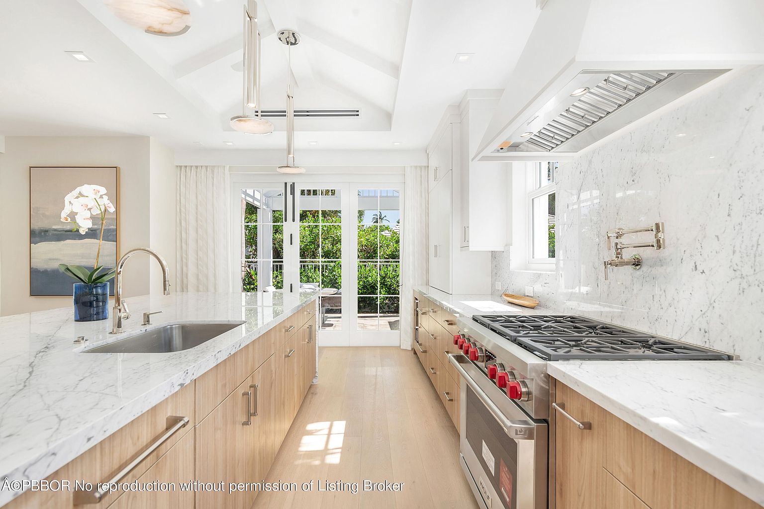 This is a bright and airy kitchen featuring a large island with a sink, marble countertops, and light wood cabinetry. A professional-grade gas range with a stainless steel hood is visible along the back wall, complemented by white marble backsplash and white cabinetry. The kitchen opens to an outdoor area through glass doors, enhancing the sense of space and light.