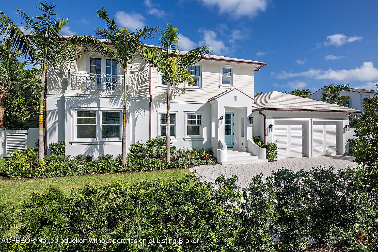 This is a front exterior view of a two-story white house with a light blue front door and a two-car garage. The house is surrounded by lush green landscaping, including palm trees and manicured bushes. The driveway is paved with light-colored stones, and the overall impression is one of luxury and curb appeal.