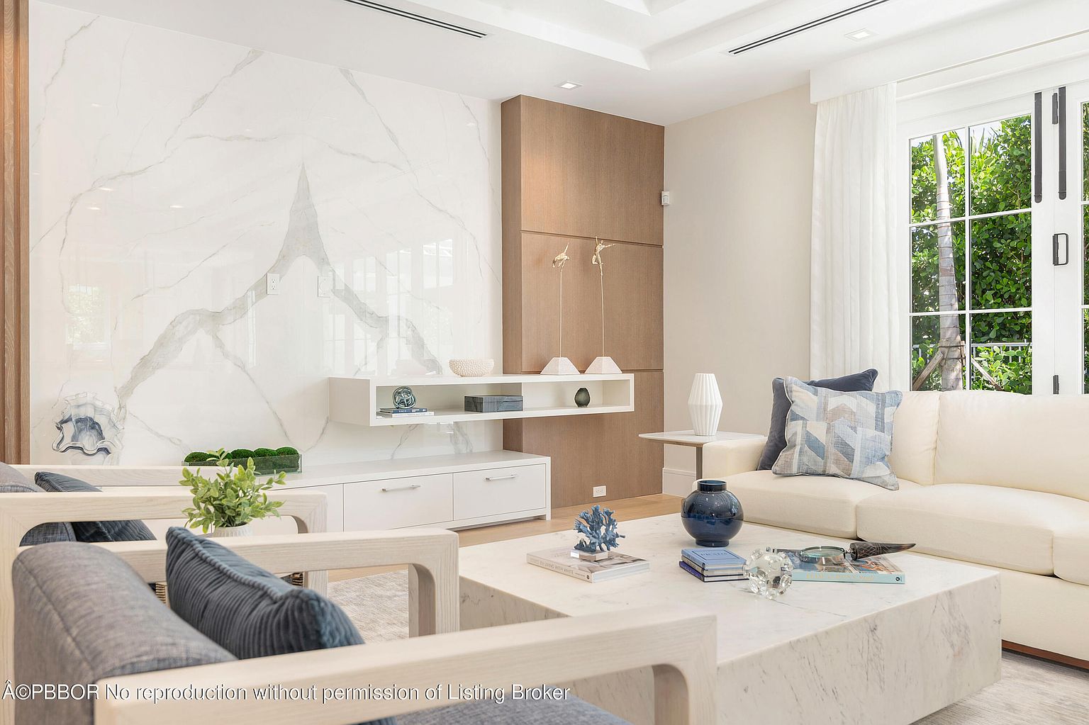 This is a bright and modern living room featuring a large marble accent wall and a built-in media center. The room is furnished with a white sofa, light wood frame chairs with blue cushions, and a marble coffee table. Large windows provide natural light and a view of the greenery outside, creating a serene and inviting atmosphere.