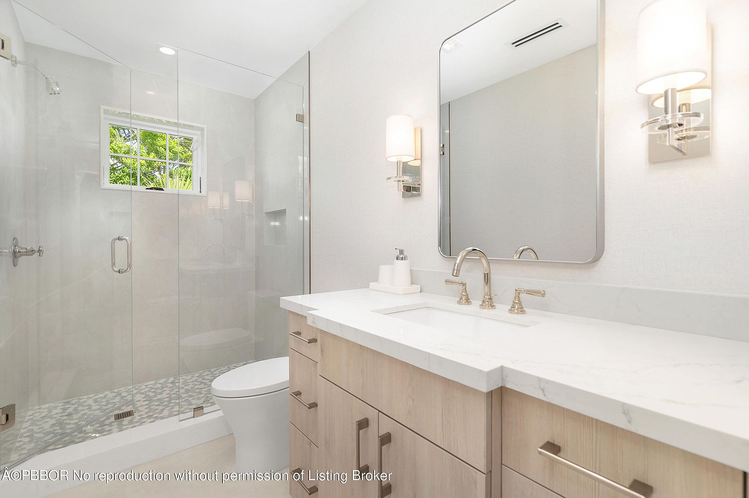 This is a bright and modern bathroom featuring a glass-enclosed shower with a mosaic tile floor, a white toilet, and a vanity with a light wood finish and white countertop. A large mirror hangs above the sink, flanked by sconce lighting, and a window provides natural light. The overall impression is clean, stylish, and well-maintained.
