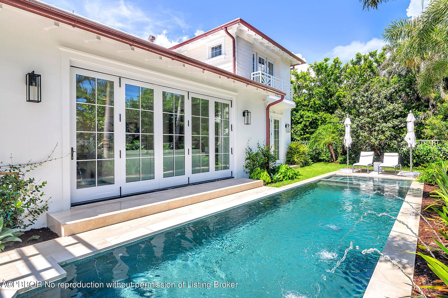 This image showcases a luxurious pool area adjacent to a modern white house. The pool features clear blue water with fountain features, surrounded by a tiled deck and lush landscaping. Lounge chairs and umbrellas are positioned near the pool, creating an inviting outdoor space perfect for relaxation and entertainment.