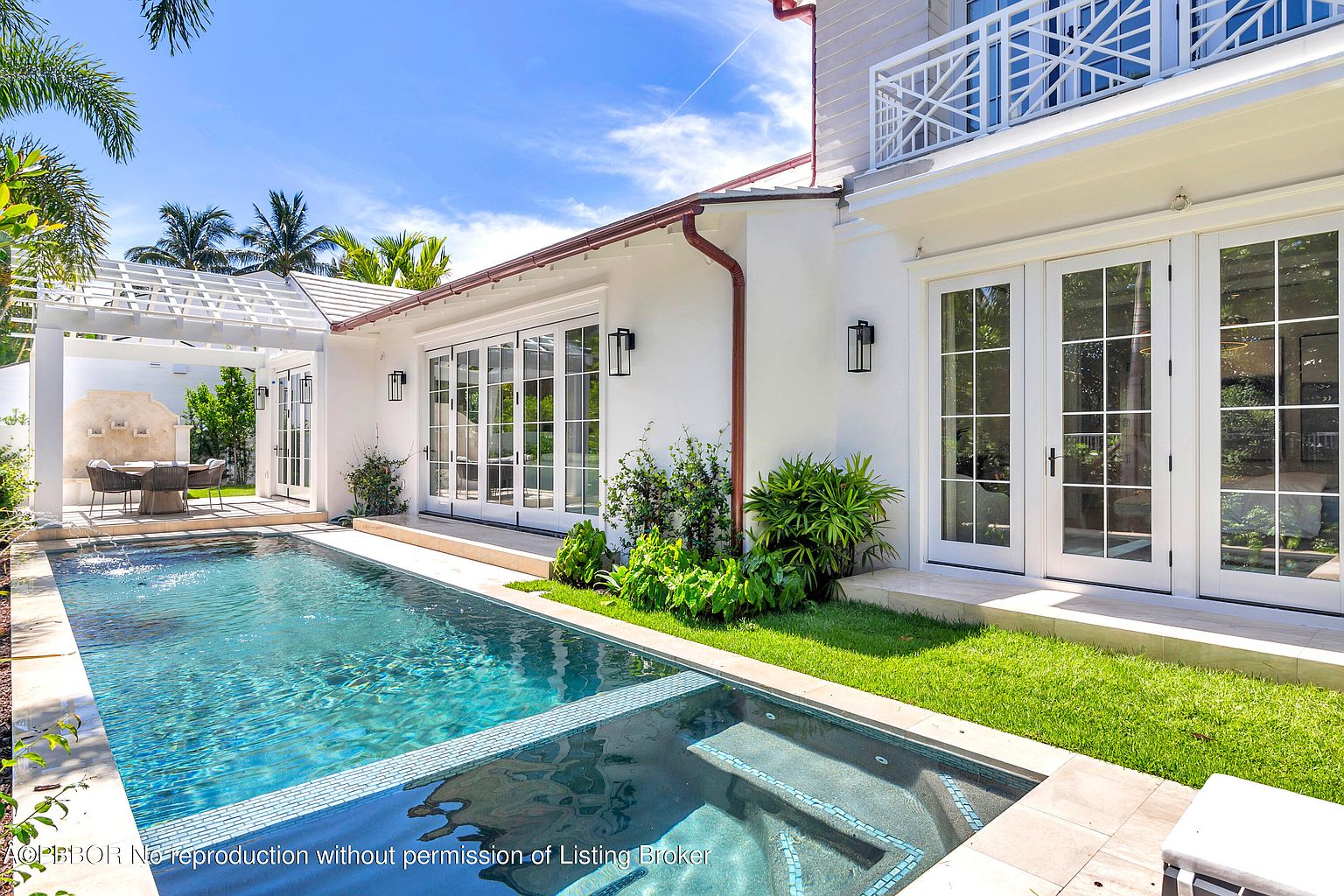 This is an exterior shot showcasing a luxurious pool and spa area of a home. The pool features clear blue water and is adjacent to a spa with tiled accents. The architecture is modern with white walls, large glass doors, and manicured landscaping, creating an inviting and upscale outdoor living space.