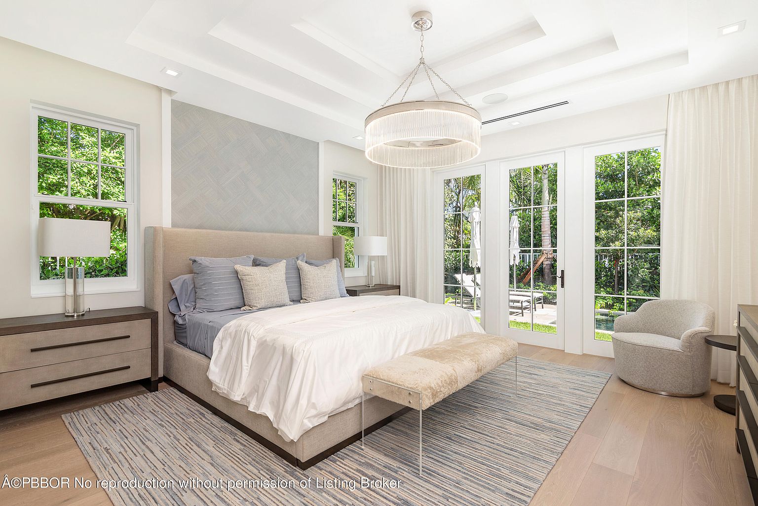 This is a bright and airy primary bedroom featuring a large bed with neutral-toned linens and pillows, flanked by matching nightstands and lamps. The room is well-lit with natural light streaming through the windows and glass doors, which lead to an outdoor area. A stylish chandelier hangs from the tray ceiling, and a soft rug adds warmth to the hardwood floors.