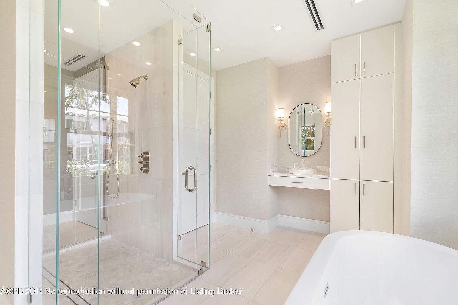 This is a bright and luxurious primary bathroom featuring a glass-enclosed shower with modern fixtures and a tiled surround. A vanity area with a round mirror and sconces is adjacent to built-in storage cabinets. The flooring is light-colored tile, and a freestanding bathtub is partially visible, contributing to the spa-like atmosphere.