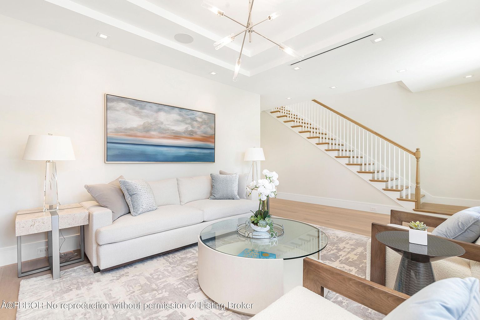 This is a bright and airy living room featuring a neutral color palette with a large painting above a comfortable sofa. A round glass coffee table sits atop a patterned rug, and a staircase is visible in the background, adding architectural interest. The room exudes a sense of calm and sophistication, perfect for relaxation and entertaining.
