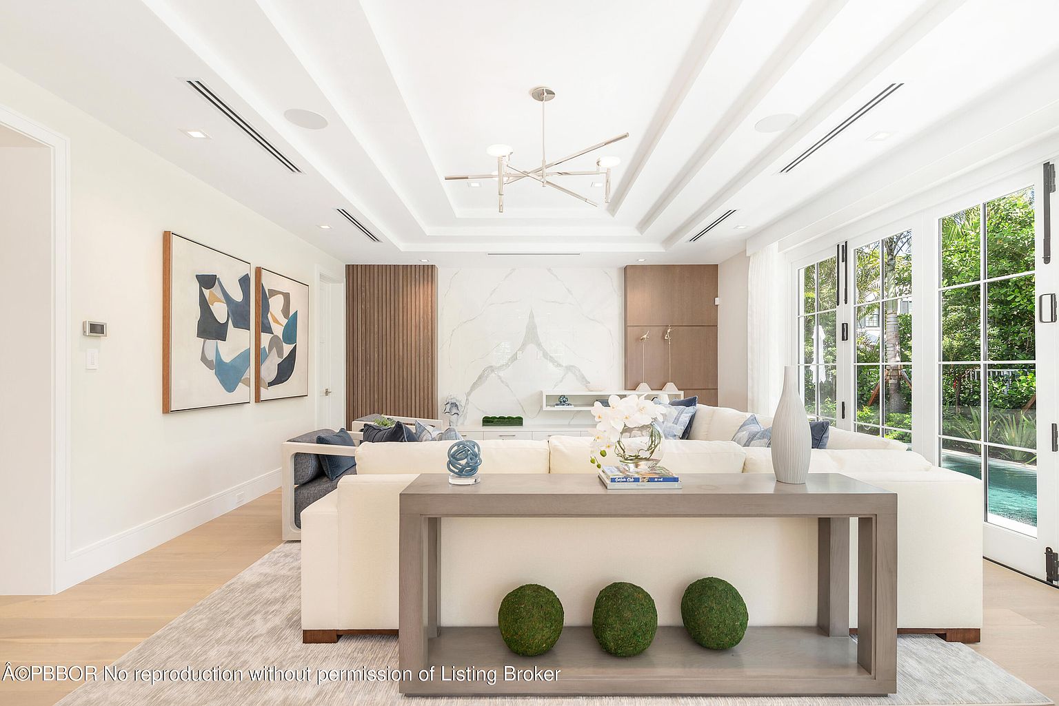 This is a bright and airy living room featuring a large, comfortable-looking sectional sofa, modern art pieces, and a stylish console table with decorative moss balls. The room is well-lit with natural light streaming through large windows and doors, complemented by modern ceiling fixtures. The overall impression is one of sophisticated comfort and contemporary design.