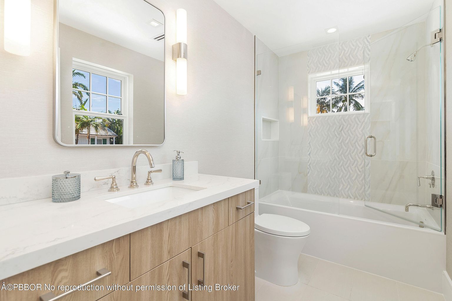 This is a bright and modern bathroom featuring a vanity with a light wood cabinet and white countertop, a rectangular mirror, and modern sconces. A toilet sits next to a bathtub with a glass shower door, and the walls are a mix of white marble and textured wallpaper. The overall impression is clean, stylish, and well-lit.