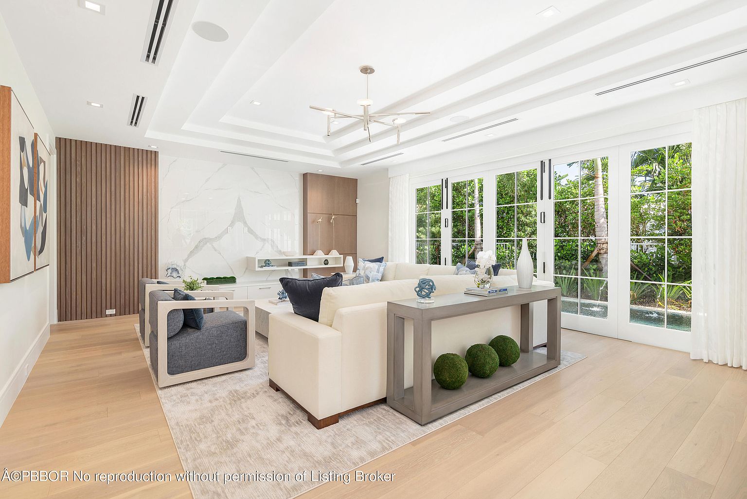 This is an interior shot of a modern living room, featuring a neutral color palette with white walls, light wood flooring, and a large area rug. The room is furnished with a white sofa, blue accent chairs, and a console table adorned with decorative items. Large windows provide ample natural light and a view of the outdoor greenery, creating a bright and airy atmosphere.
