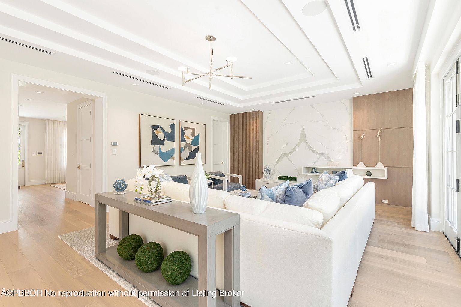 This is an interior shot of a modern living room featuring a large white sectional sofa with blue pillows. The room is decorated with abstract art, a decorative console table with moss balls, and a unique chandelier. The space is bright and airy, with light wood floors and a marble accent wall, creating a luxurious and inviting atmosphere.