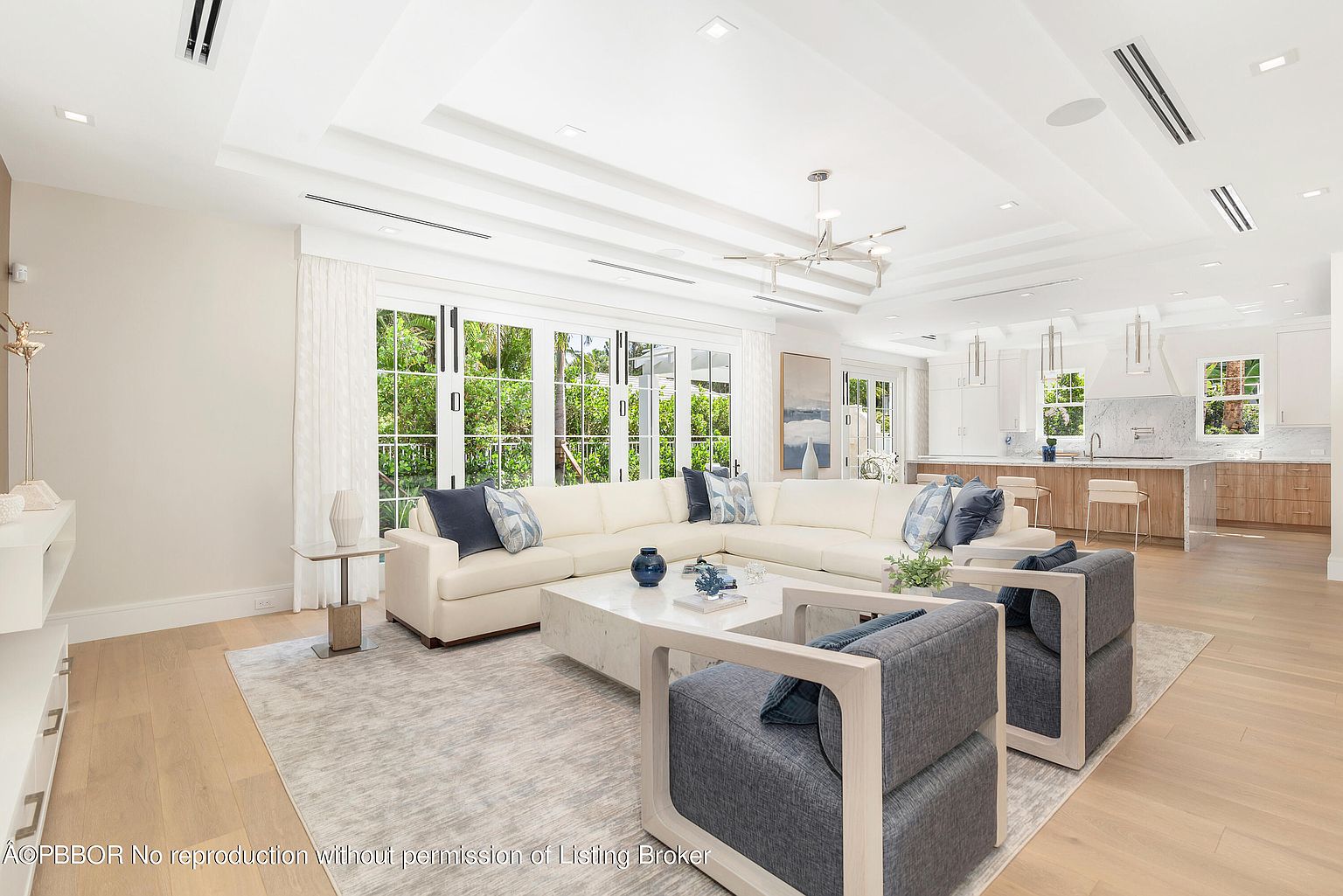 This is an interior shot of a bright and airy living room that seamlessly transitions into a modern kitchen. The living room features a large, white sectional sofa adorned with blue pillows, complemented by two gray armchairs and a marble coffee table. Large windows provide ample natural light and views of the lush greenery outside, enhancing the open and inviting atmosphere.