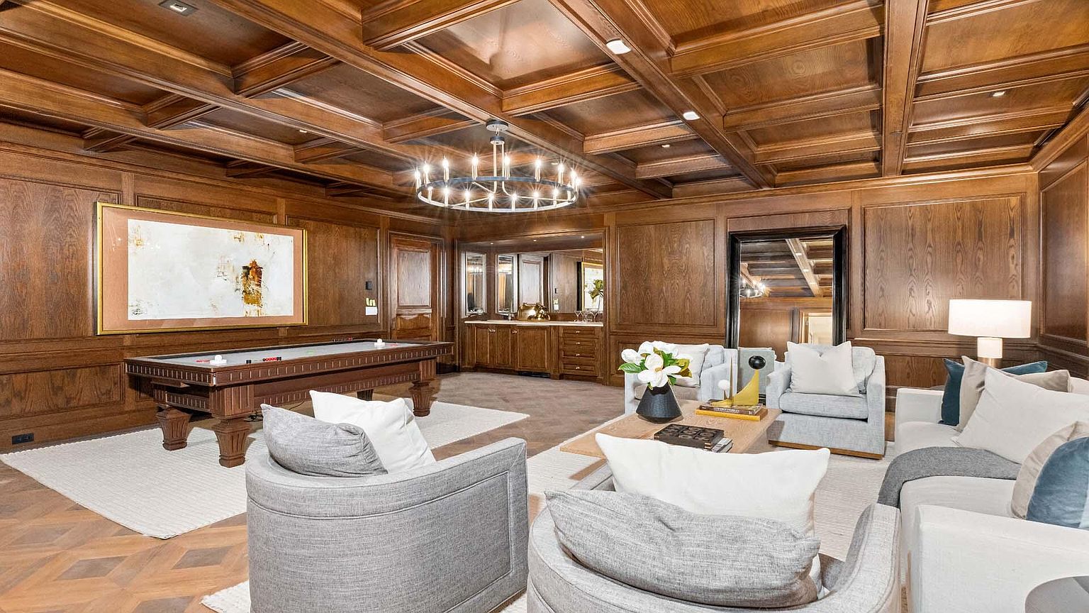 This is a luxurious living room featuring rich wood paneling and coffered ceilings, creating a warm and sophisticated ambiance. The room includes a pool table, a wet bar, and comfortable seating arrangements with plush armchairs and a sofa, all centered around a stylish coffee table. The lighting is provided by an elegant chandelier, enhancing the room's upscale feel.