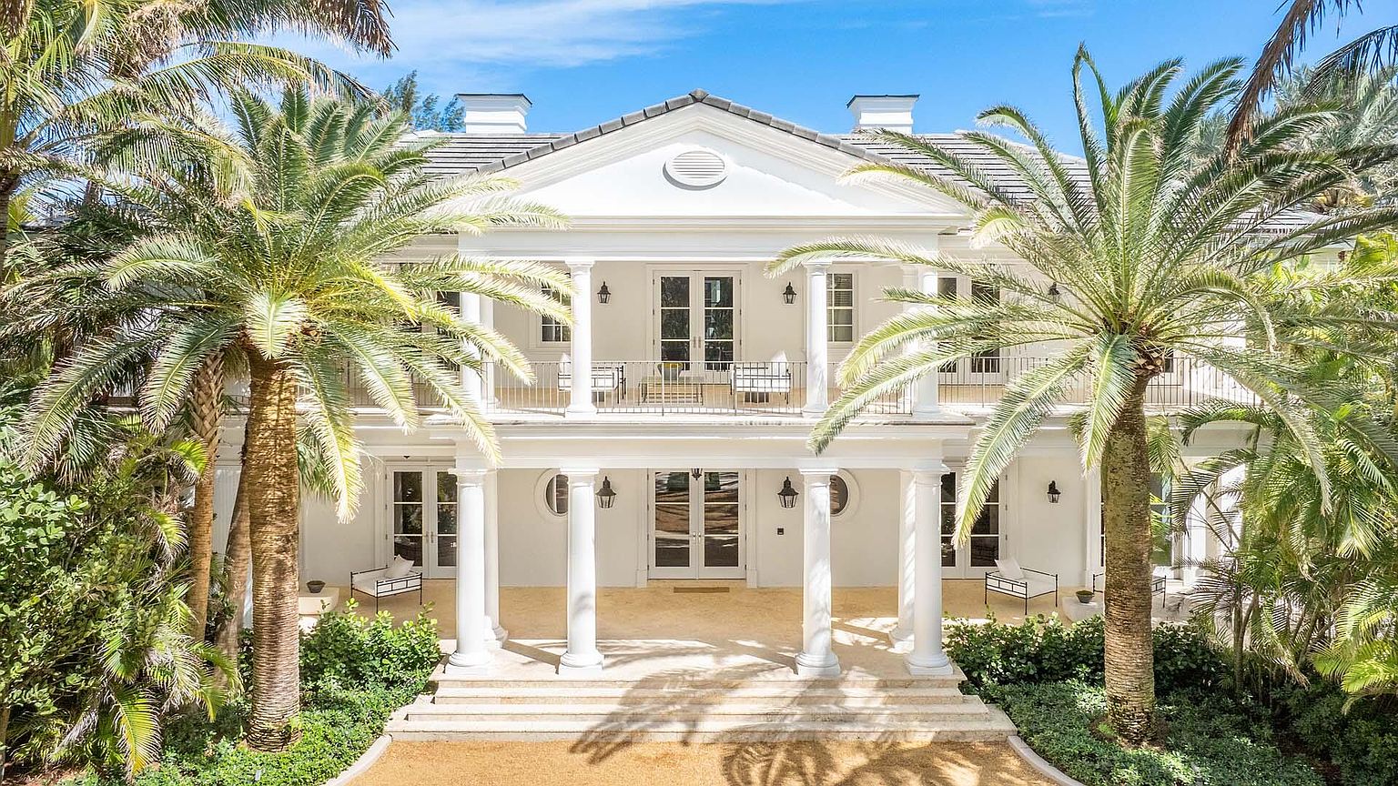 This is a front view of a grand, two-story white house with a symmetrical facade. The house features a prominent portico supported by white columns, a second-story balcony, and a well-manicured lawn. Palm trees frame the house, adding a tropical feel to the property.