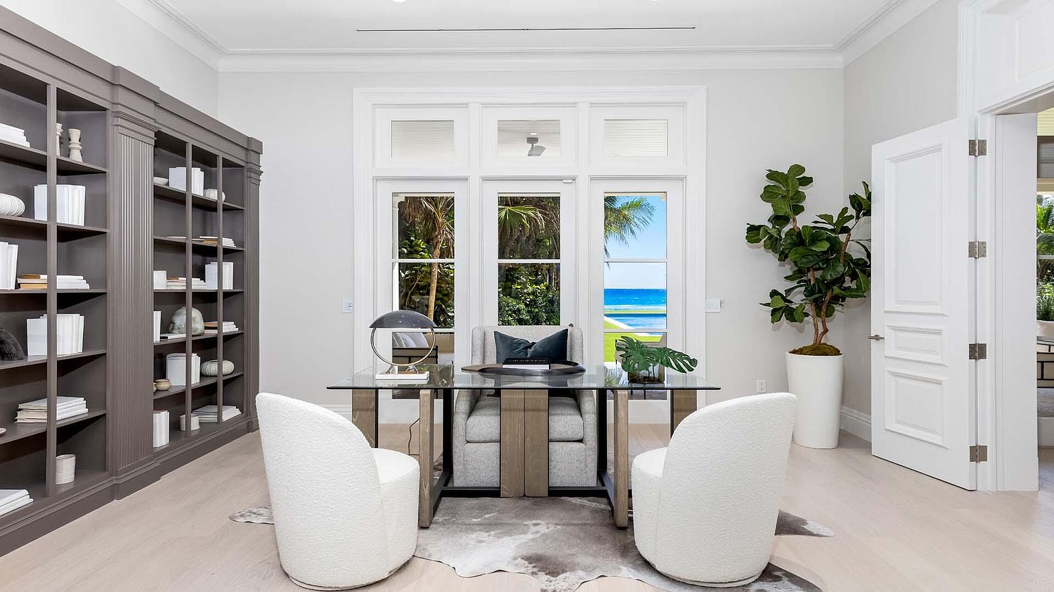 This is an elegant home office featuring a glass-top desk with two white chairs, a large window with a view of the ocean, and a built-in bookshelf. The room is decorated in neutral tones, creating a calm and sophisticated atmosphere. A large potted plant adds a touch of nature to the space.