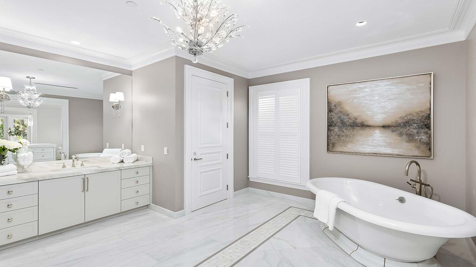 This is a luxurious primary bathroom featuring a freestanding white bathtub with a chrome faucet, a white vanity with marble countertops, and a large mirror. The room is decorated in neutral tones with white trim and a decorative chandelier, creating a serene and elegant atmosphere. A large painting hangs on the wall above the tub, and a window with white shutters provides natural light.