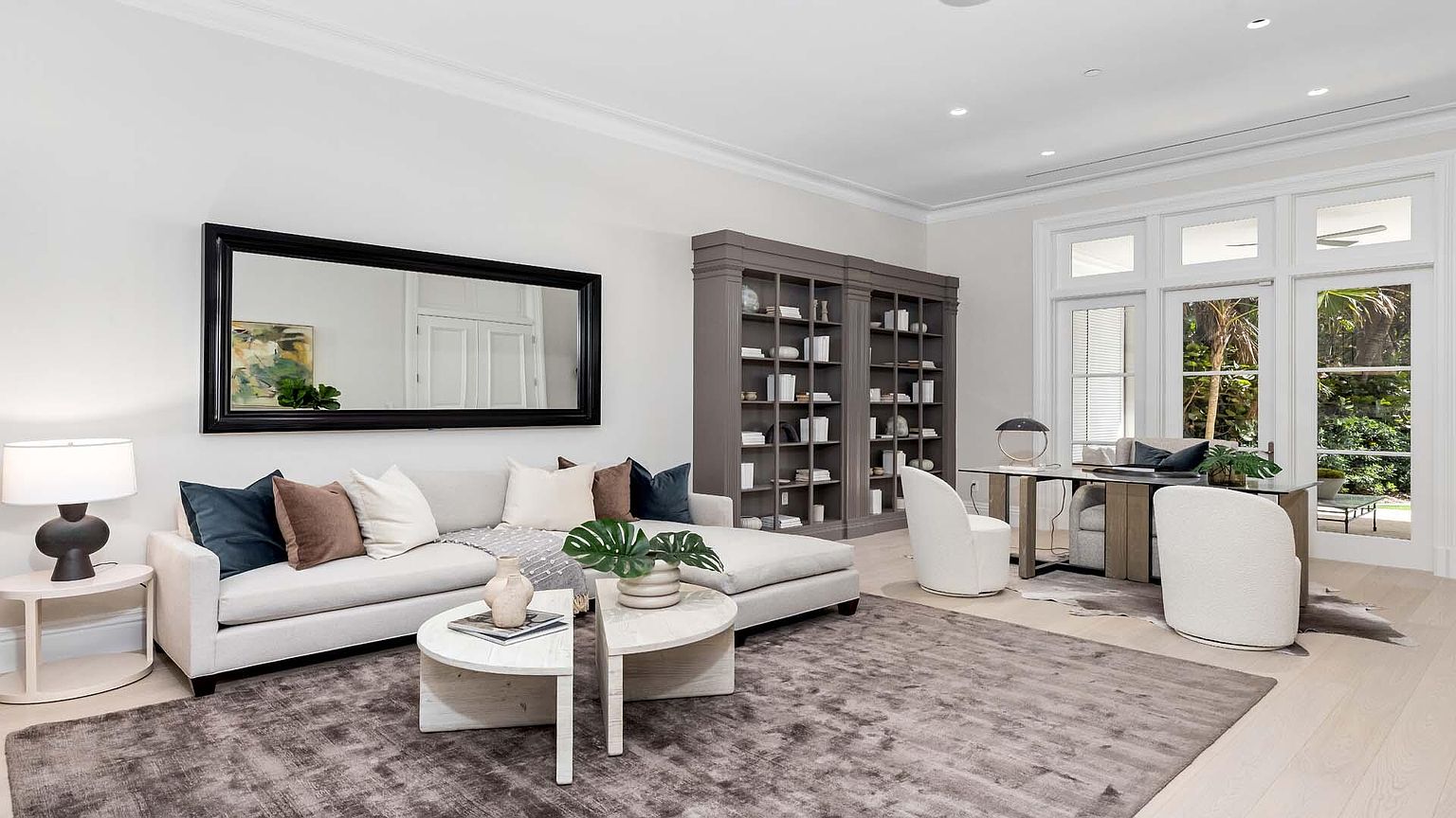This is an interior shot of a living room featuring a neutral color palette. The room includes a sectional sofa with decorative pillows, a round coffee table, and a large mirror above the sofa. A bookshelf is placed against the wall, and a desk with chairs is positioned near the windows, creating a combined living and office space.