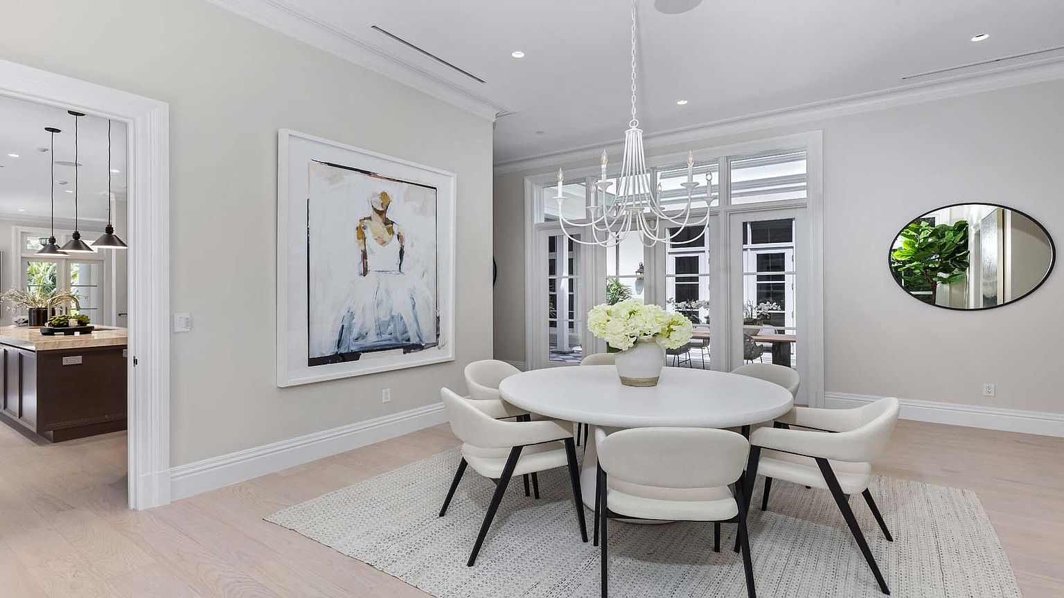This is an interior shot of a dining room featuring a round white table surrounded by six chairs with black legs. A modern white chandelier hangs above the table, and a large framed painting adorns the wall. The room is bright and airy, with natural light streaming in through the French doors leading to an outdoor area, and a round mirror with a plant is on the right wall.
